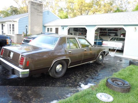 2001 Ford Crown Victoria - A brown sedan is parked in a driveway next to a garage with other vehicles visible inside.