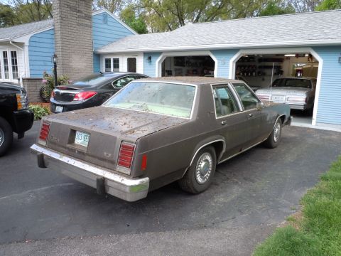 2001 Ford Crown Victoria - A brown 1980s Ford sedan is parked in front of a garage, with another car visible inside.