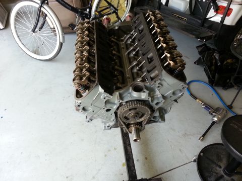 1994 Ford Bronco - Engine assembly on a stand, showing the timing chain and gears. A bicycle and golf cart are partially visible in the background.