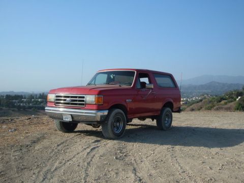 1988 Ford Bronco - A red Ford Bronco is parked on a dirt road overlooking a town and hills under a clear blue sky.