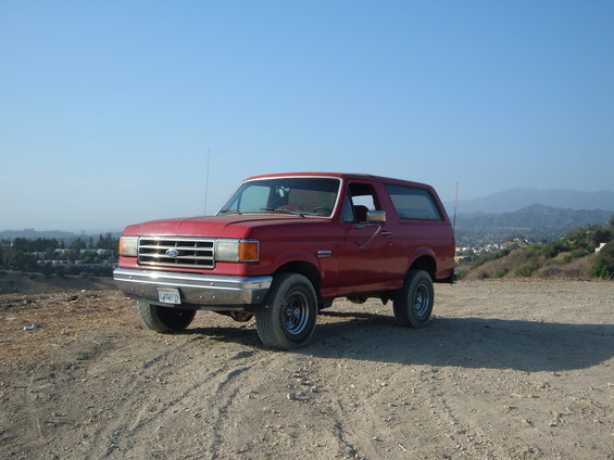 Red Ford Bronco parked on a dirt road with a town and hills in the background