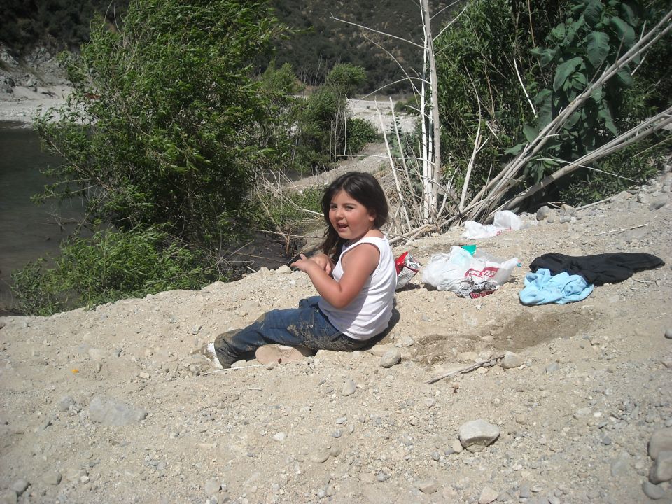 Girl in white tank top and jeans sits on dirt next to a river with trash nearby