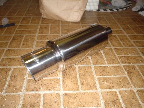 1999 Ford Taurus - A brand-new polished stainless steel canister muffler with an angle-cut rolled tip and small inlet pipe, lying on a tile floor next to its…