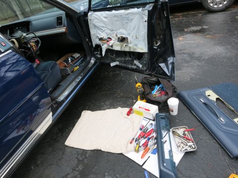 1991 Ford LTD Crown Victoria - The interior of a dark blue car is visible with the driver's side door open and the door panel removed, revealing tools and parts on the gr…
