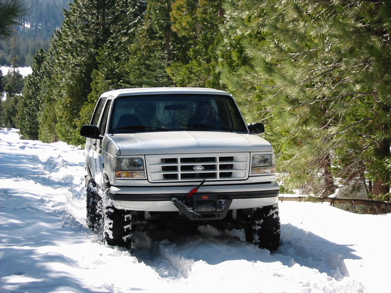 White Ford Bronco with Warn winch parked in deep snow on pine-lined forest road