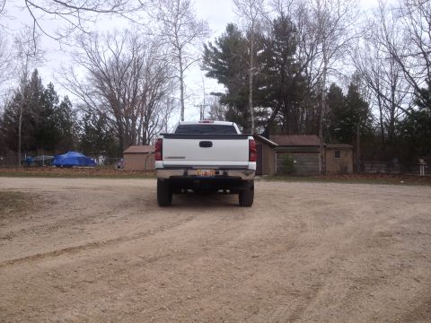 2006 Chevrolet Silverado 2500 - A white 2006 Chevrolet Silverado 2500 pickup truck is seen from the rear in an outdoor setting with trees and buildings in the background.