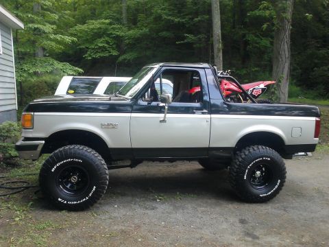 1991 Ford Bronco - This lifted Ford Bronco is parked next to a house with trees in the background.