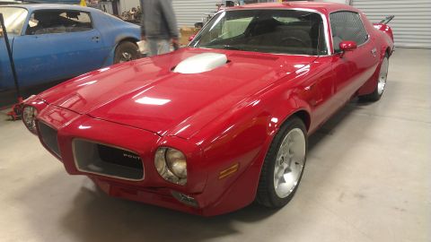 1972 Pontiac Trans Am - A red 1970s Pontiac Firebird is parked next to a blue car in a garage. The red car has a prominent shaker hood scoop and custom wheels.