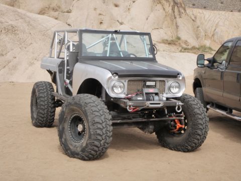 1961 International Scout 800 - A heavily modified off-road vehicle with a roll cage sits in a sandy, rocky area next to a dark pickup truck.