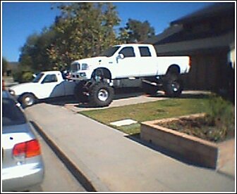1992 Ford Bronco - A heavily modified white pickup truck dominates the scene, dwarfing a smaller white vehicle parked nearby.
