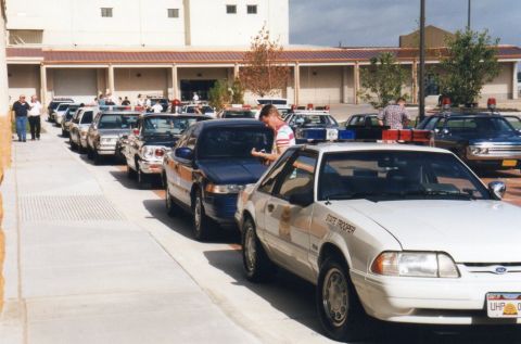 1991 Ford LTD Crown Victoria - A line of police cars, including a white 1991 Ford Crown Victoria, are parked outside a building.