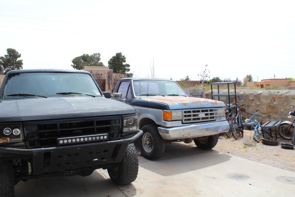 Two trucks parked outside, one black with a light bar, the other blue and silver with rust. Bicycles and a motorcycle are nearby