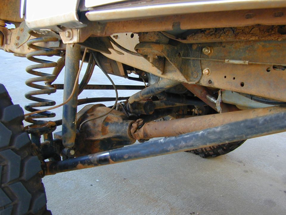 Rusty undercarriage of a Bronco showing front axle, coil spring, and driveshaft over concrete.
