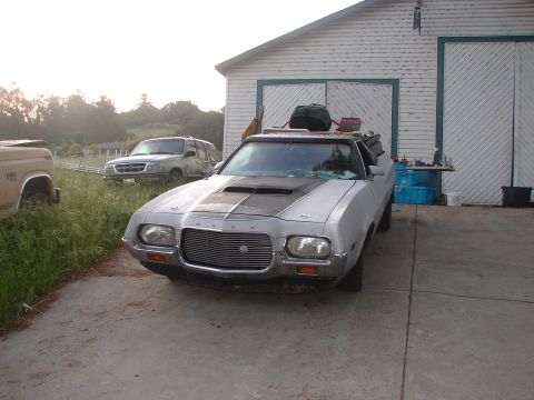 1973 Ford Ranchero - Ford Ranchero with hood scoop and luggage on roof parked outside a garage, with other vehicles nearby.