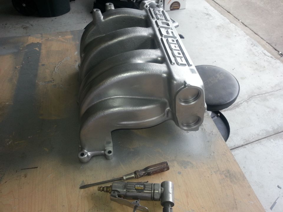 Silver aluminum engine intake manifold with 'FORD' embossed on top, resting on a wooden surface with tools