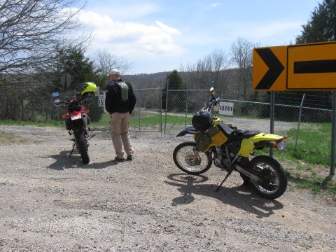 2006 Suzuki DR-Z400S - Two dual-sport motorcycles are parked on a gravel lot next to a fence with a yellow directional sign overhead.
