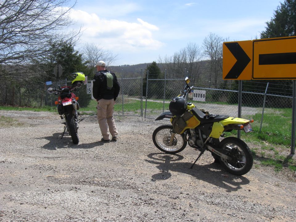 Two dirt bikes parked on a gravel lot near a fence and a yellow directional sign. A person stands between them