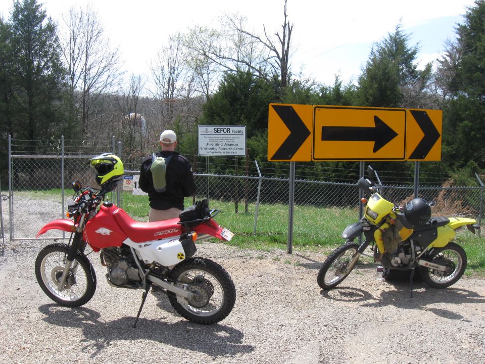 Red and yellow dirt bikes parked on gravel near a fence and directional signs