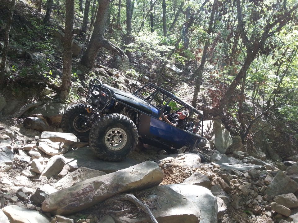 Blue off-road buggy with roll cage navigating a rocky, wooded trail