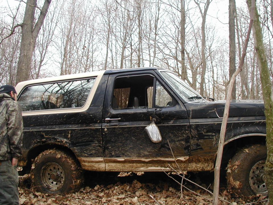 Mud-splattered black Ford Bronco with tan roof, side view in bare winter woods