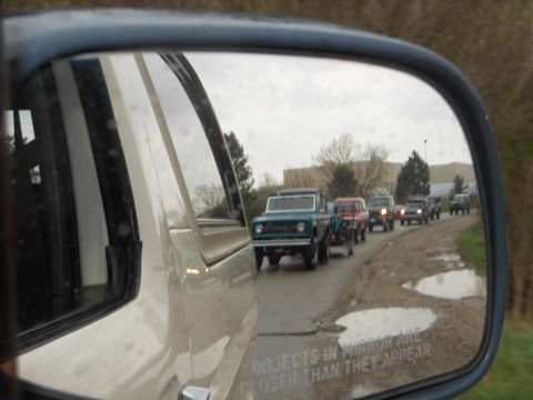 1995 Ford Bronco - A convoy of classic Ford Broncos drives down a wet, unpaved road, reflected in a side mirror.
