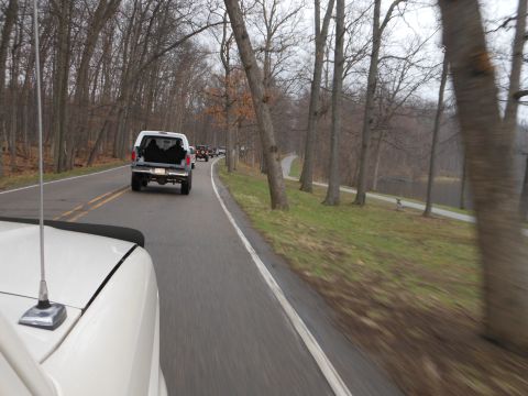 1995 Ford Bronco - A white 1995 Ford Bronco leads a line of vehicles on a road lined with bare trees.