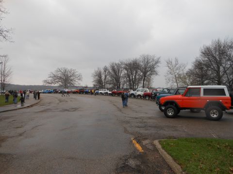 1995 Ford Bronco - A line of vehicles, including a prominent orange first-generation Bronco, are parked outdoors on a cloudy day.