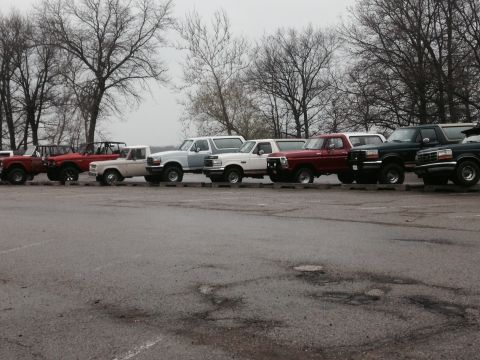 1995 Ford Bronco - A row of Ford Broncos and F-Series trucks are parked side-by-side under a cloudy sky.