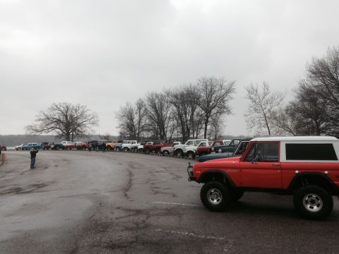 1995 Ford Bronco - A group of Ford Broncos are gathered in a parking lot near a body of water on an overcast day.