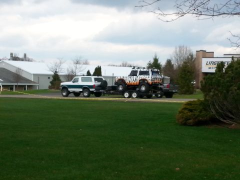 1995 Ford Bronco - A white and green Bronco tows a trailer carrying a custom-painted Bronco, with a building in the background.