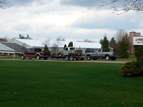 1995 Ford Bronco - A convoy of three trucks, one towing a trailer with another truck, moves past a building.