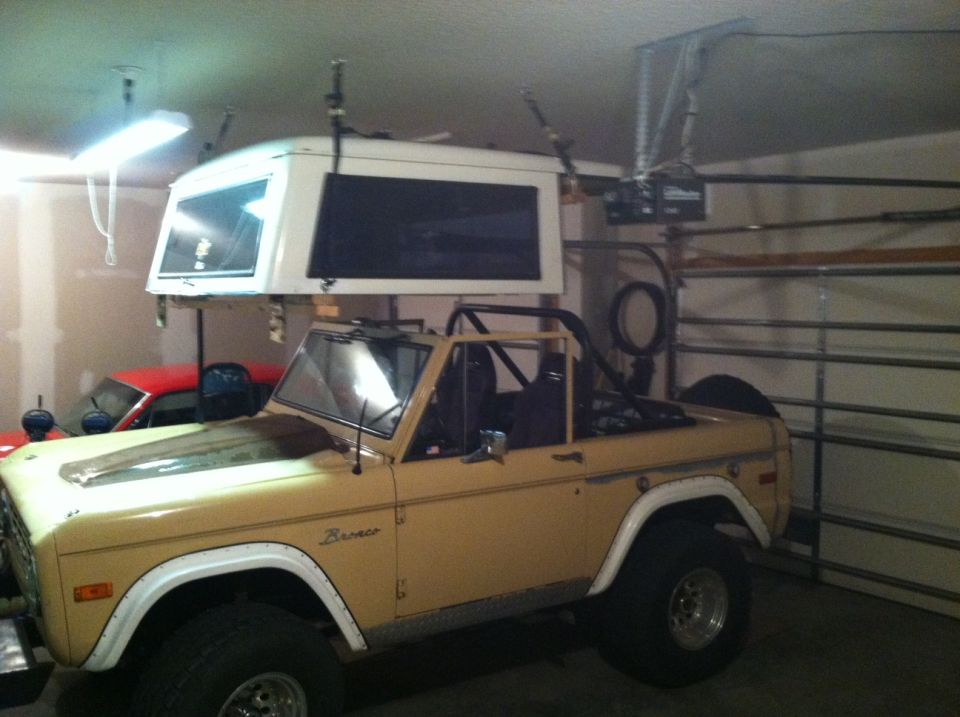 1976 Ford Bronco in a garage with a white camper shell suspended above it