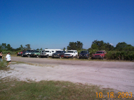Row of parked trucks and SUVs lined up at edge of a dirt clearing under blue sky