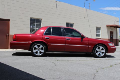 2002 Mercury Grand Marquis - Side view of a red 2002 Mercury Grand Marquis parked in front of a beige building.