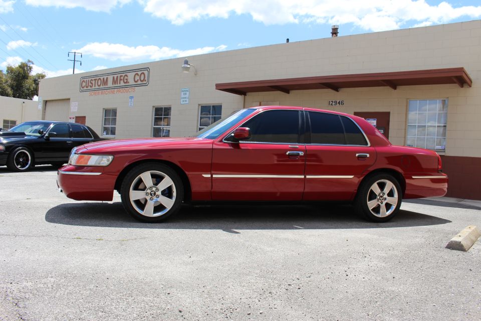 Red sedan parked outside a building with 'CUSTOM MFG. CO.' sign. Black sedan behind it