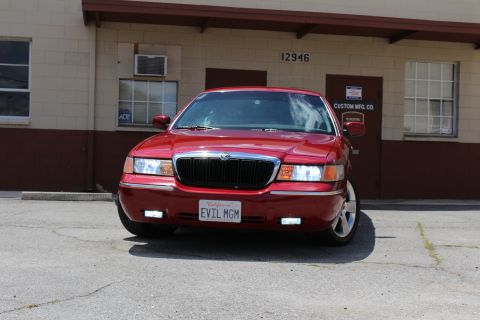 2002 Mercury Grand Marquis - A red 2002 Mercury Grand Marquis is parked in front of a building, displaying a custom "EVIL MGM" license plate.