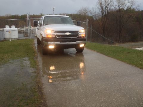 2006 Chevrolet Silverado 2500 - A white pickup truck is parked on a wet road with its headlights illuminated.
