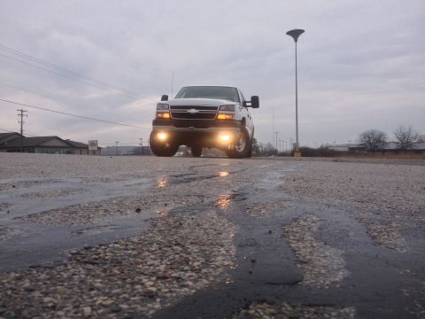 2006 Chevrolet Silverado 2500 - A white 2006 Chevrolet Silverado 2500 truck is seen from a low angle on a wet road with its headlights illuminated.