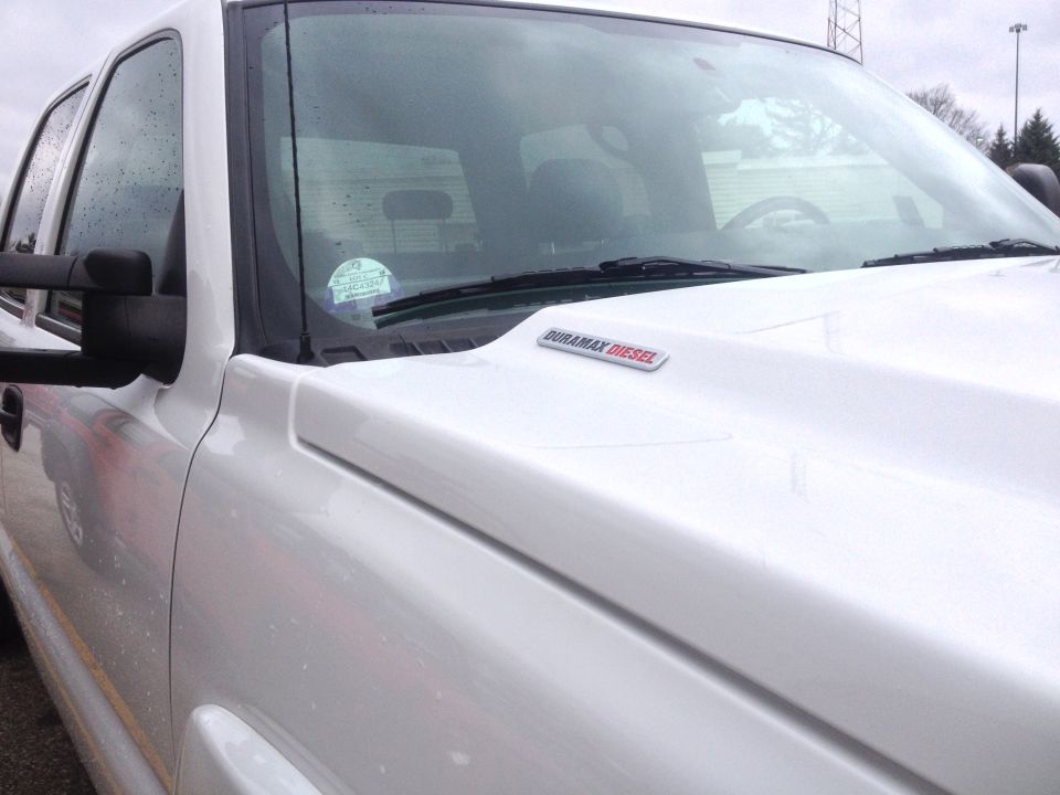 White pickup truck, hood has a Duramax Diesel badge. Windshield has a sticker