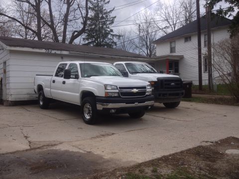 2006 Chevrolet Silverado 2500 - Two white pickup trucks are parked in a driveway in front of a house and garage.