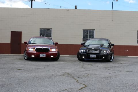 2002 Mercury Grand Marquis - Two sedans, a red 2002 Mercury Grand Marquis and a black modified vehicle, are parked side-by-side in front of a brick building.
