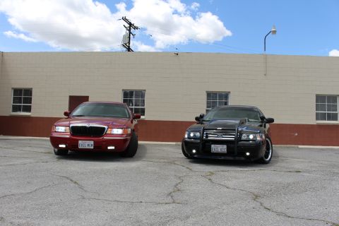 2002 Mercury Grand Marquis - Two sedans, one red and one black with a push bar, are parked in front of a beige building under a cloudy sky.