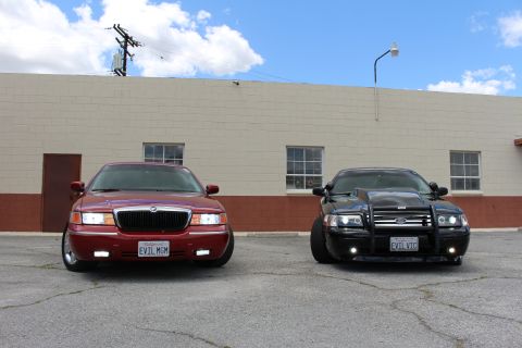 2002 Mercury Grand Marquis - Two cars, a red Mercury Grand Marquis and a black modified vehicle, are parked side-by-side in front of a beige building.