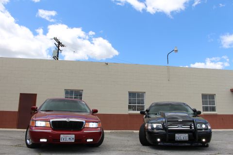 2002 Mercury Grand Marquis - Two sedans, one red and one black, are parked in front of a light-colored building under a blue sky with clouds.