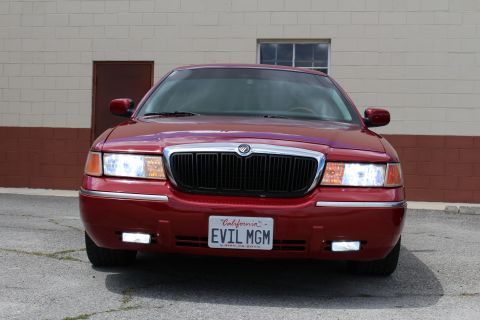 2002 Mercury Grand Marquis - A red 2002 Mercury Grand Marquis is parked in front of a brick building, displaying its front grille and headlights.
