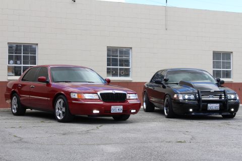 2002 Mercury Grand Marquis - Two sedans, one red and one black with a modified hood and push bar, are parked in front of a building.