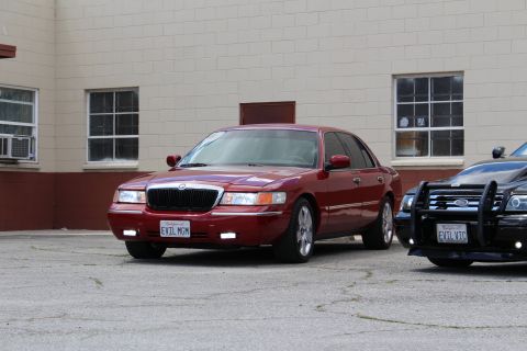 2002 Mercury Grand Marquis - A red 2002 Mercury Grand Marquis is parked in front of a building, with a black police car to its right.