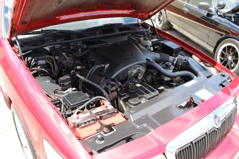 2002 Mercury Grand Marquis - Engine bay of a red 2002 Mercury Grand Marquis, showing the V8 engine and battery. Another car is visible in the background.