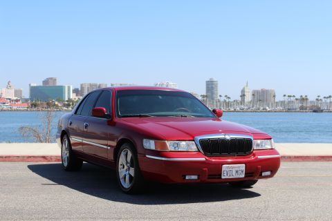 2002 Mercury Grand Marquis - A red 2002 Mercury Grand Marquis is parked near a waterfront with a city skyline in the distance.