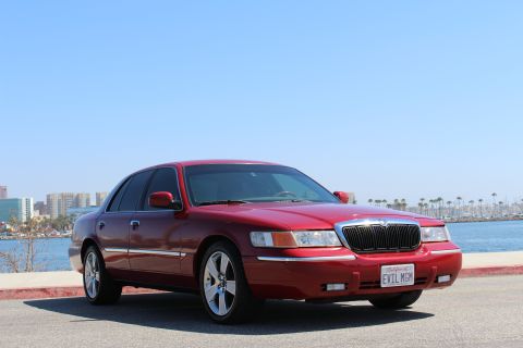 2002 Mercury Grand Marquis - A red 2002 Mercury Grand Marquis is parked near a body of water with a city skyline in the background.